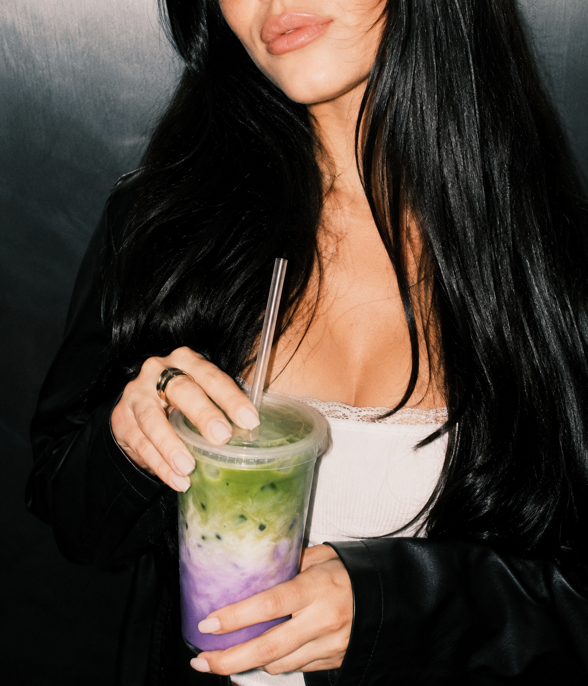 A woman in a black jacket holding a layered green matcha and purple ube iced drink.