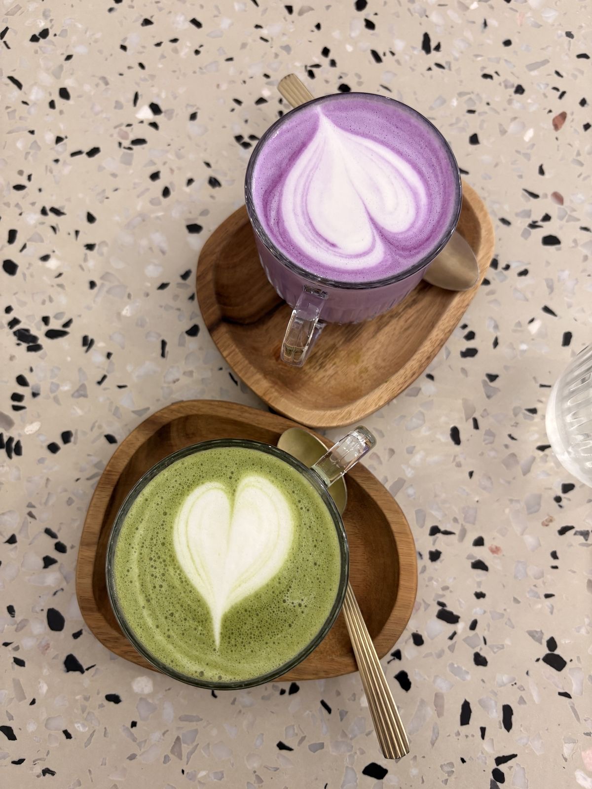A purple ube latte and a matcha latte side by side on a terrazzo table, both with heart latte art.